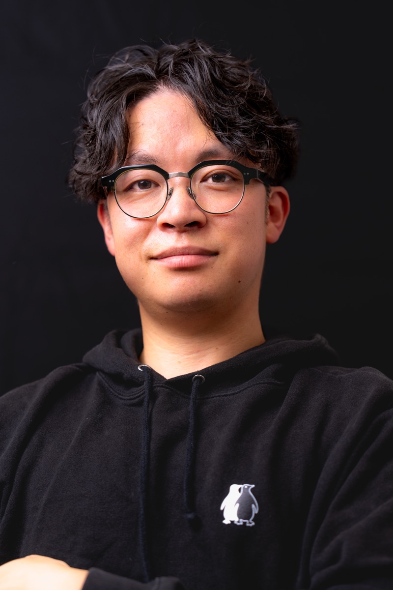黒背景に浮かぶ代表・佐藤明彦のポートレート。眼鏡をかけ、静かにこちらを見ている。